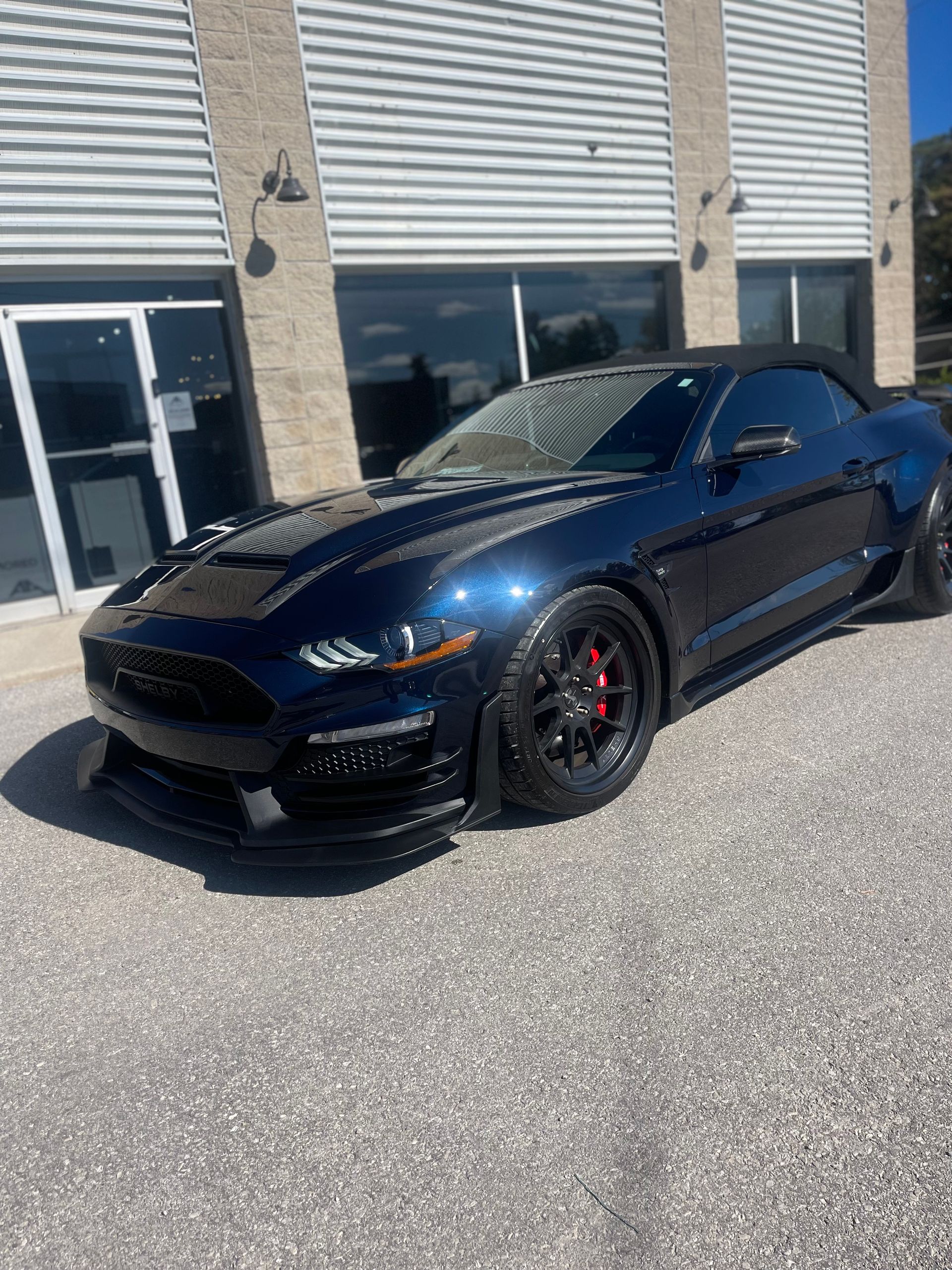 A dark blue Ford Mustang convertible parked in front of a building with light-colored siding and glass doors.