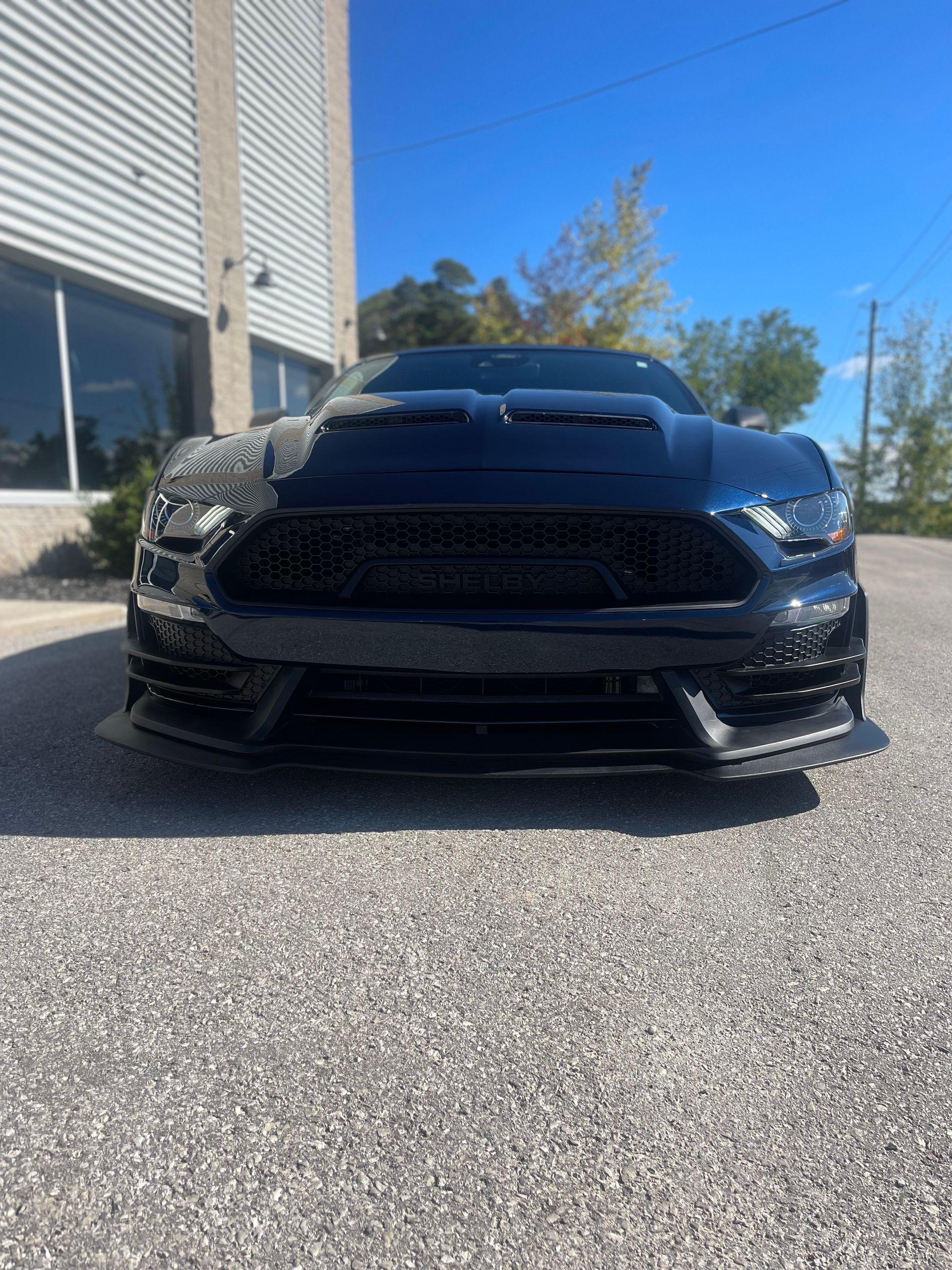 A front-facing view of a dark-colored Ford Mustang parked on a gravel lot next to a building on a sunny day.