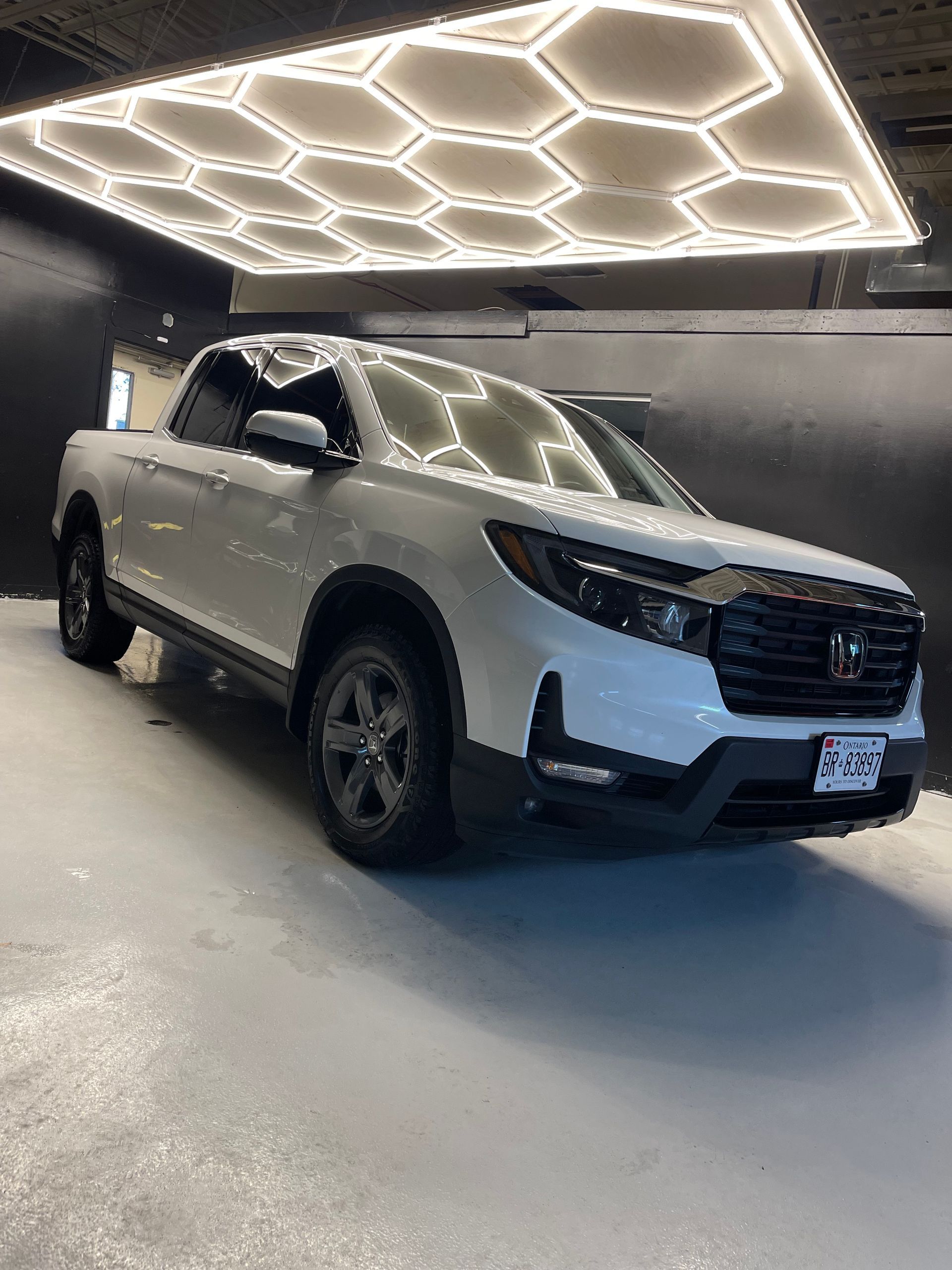 A white Honda Ridgeline pickup truck parked in an indoor garage under hexagonal LED ceiling lights.