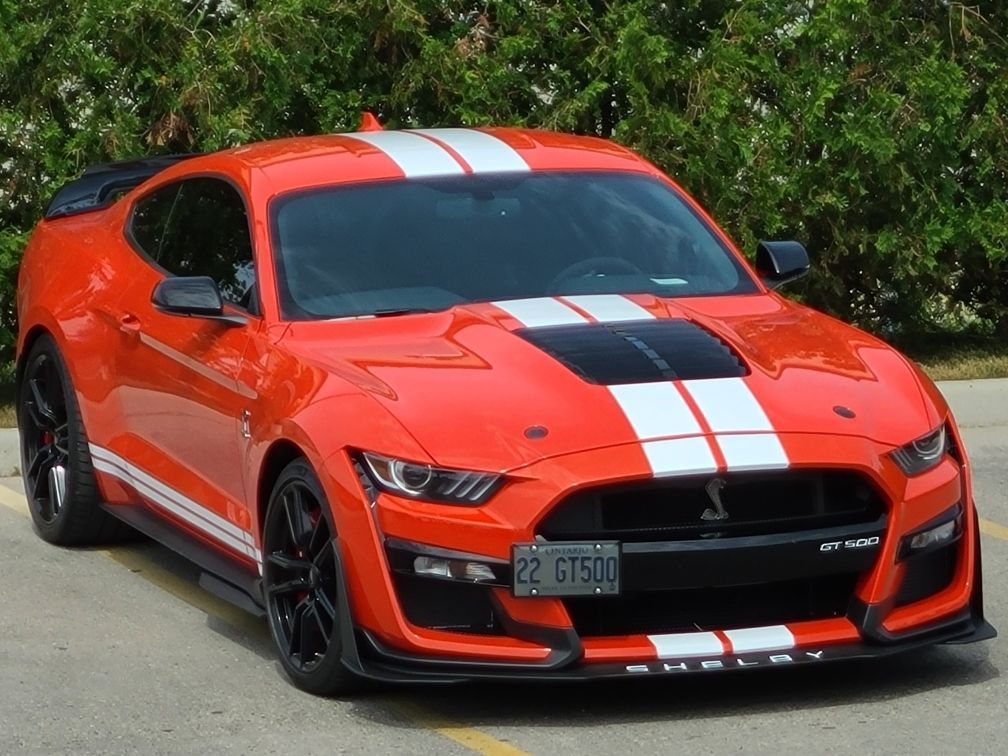 An orange Ford Mustang Shelby GT500 with white racing stripes parked on asphalt.