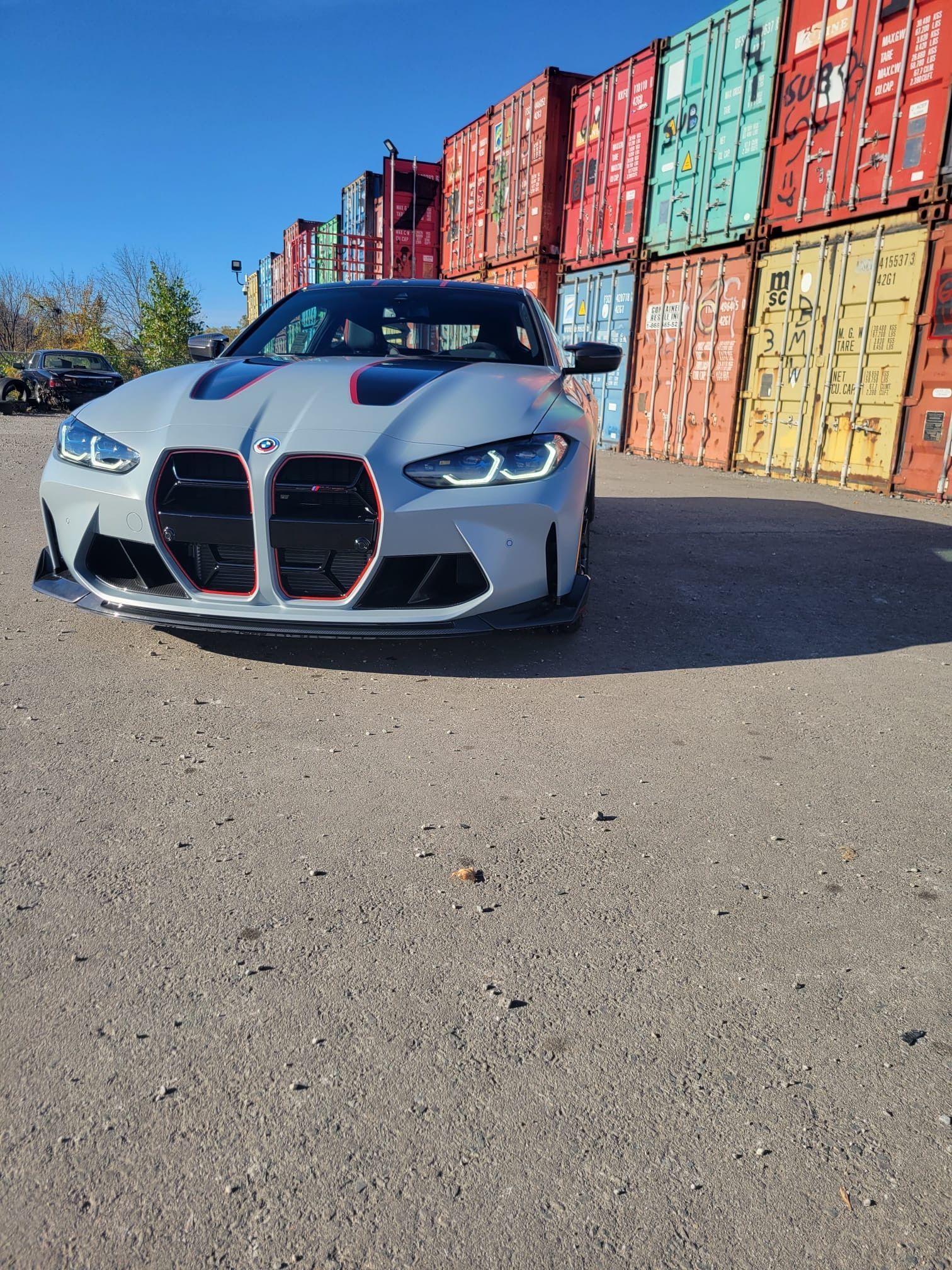 A grey BMW M4 sports car with red accents parked in front of stacked, multicolored shipping containers.