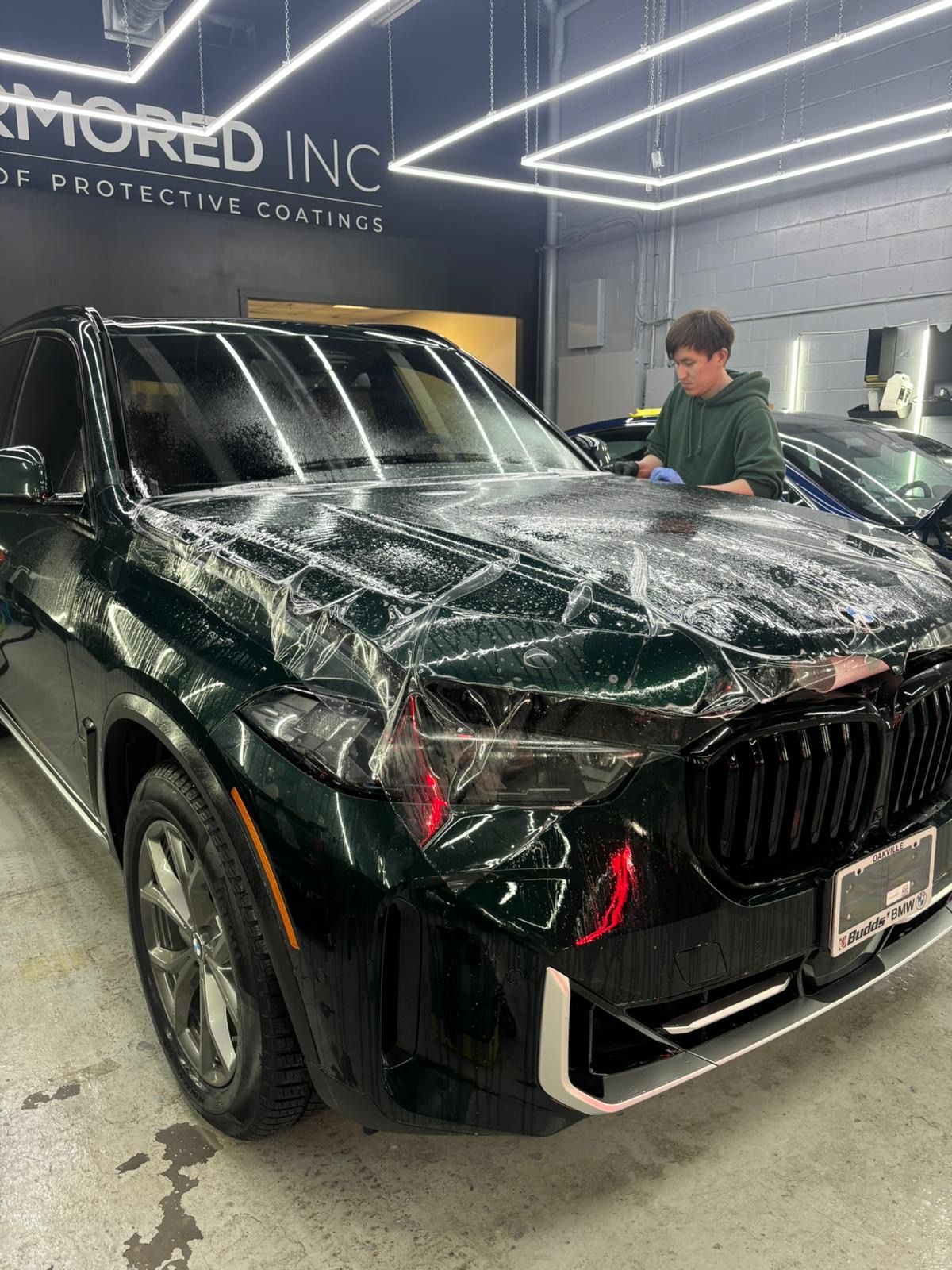 A technician applies a clear protective film to the hood of a dark green BMW SUV inside a professional detailing shop.