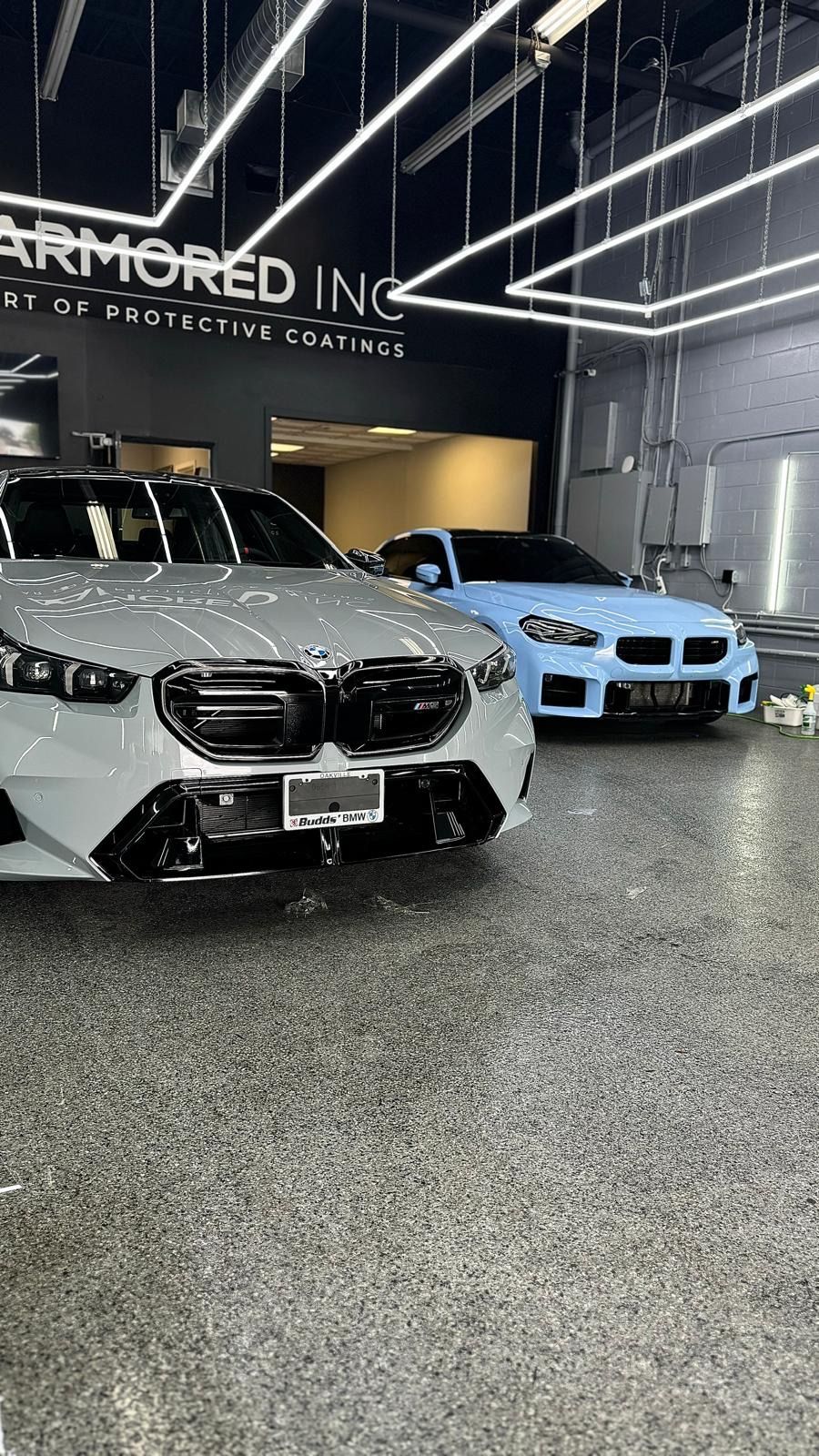Two BMWs parked in a professional detailing studio with neon lighting and speckled flooring.