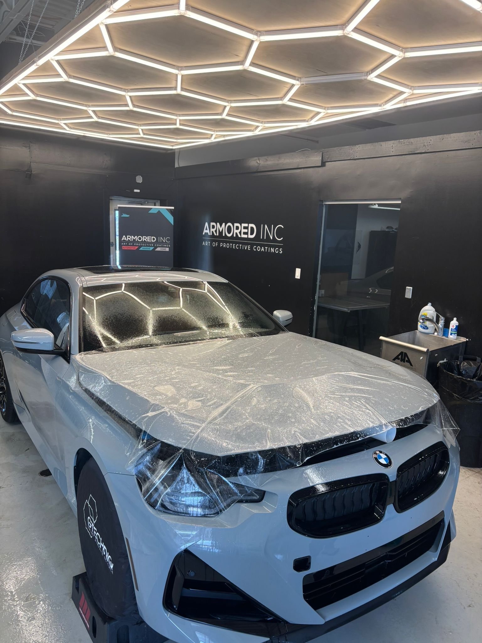 A gray BMW in a shop with black walls and hexagonal ceiling lights, undergoing a clear paint protection film application.