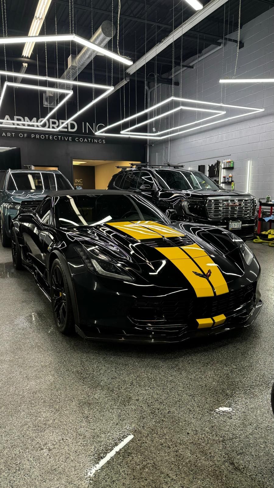 A black Corvette with a wide yellow racing stripe parked inside a modern showroom with overhead geometric lighting.