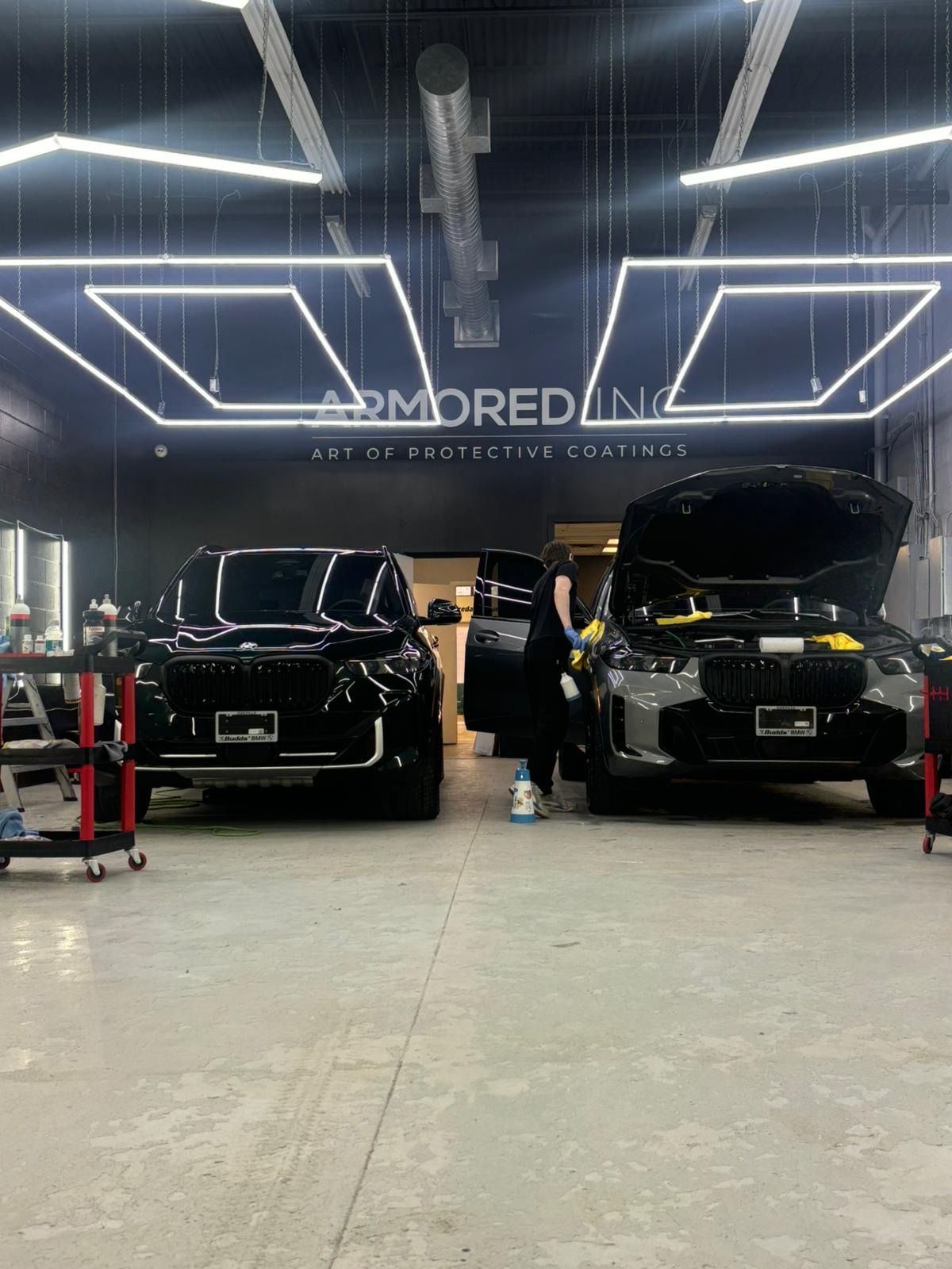 A technician works on a black vehicle in a brightly lit automotive detailing shop with another car parked nearby.