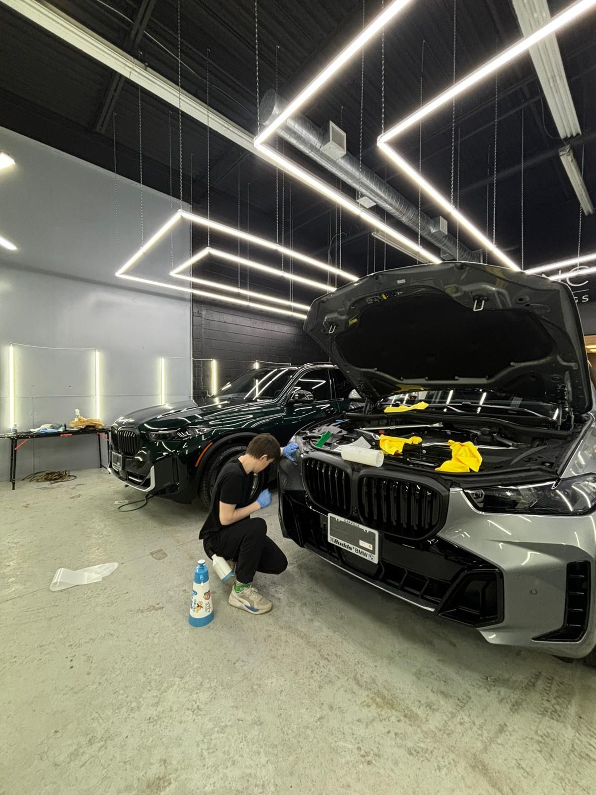 A person in gloves cleaning the front of a gray BMW in a bright, modern garage with another dark car parked behind.