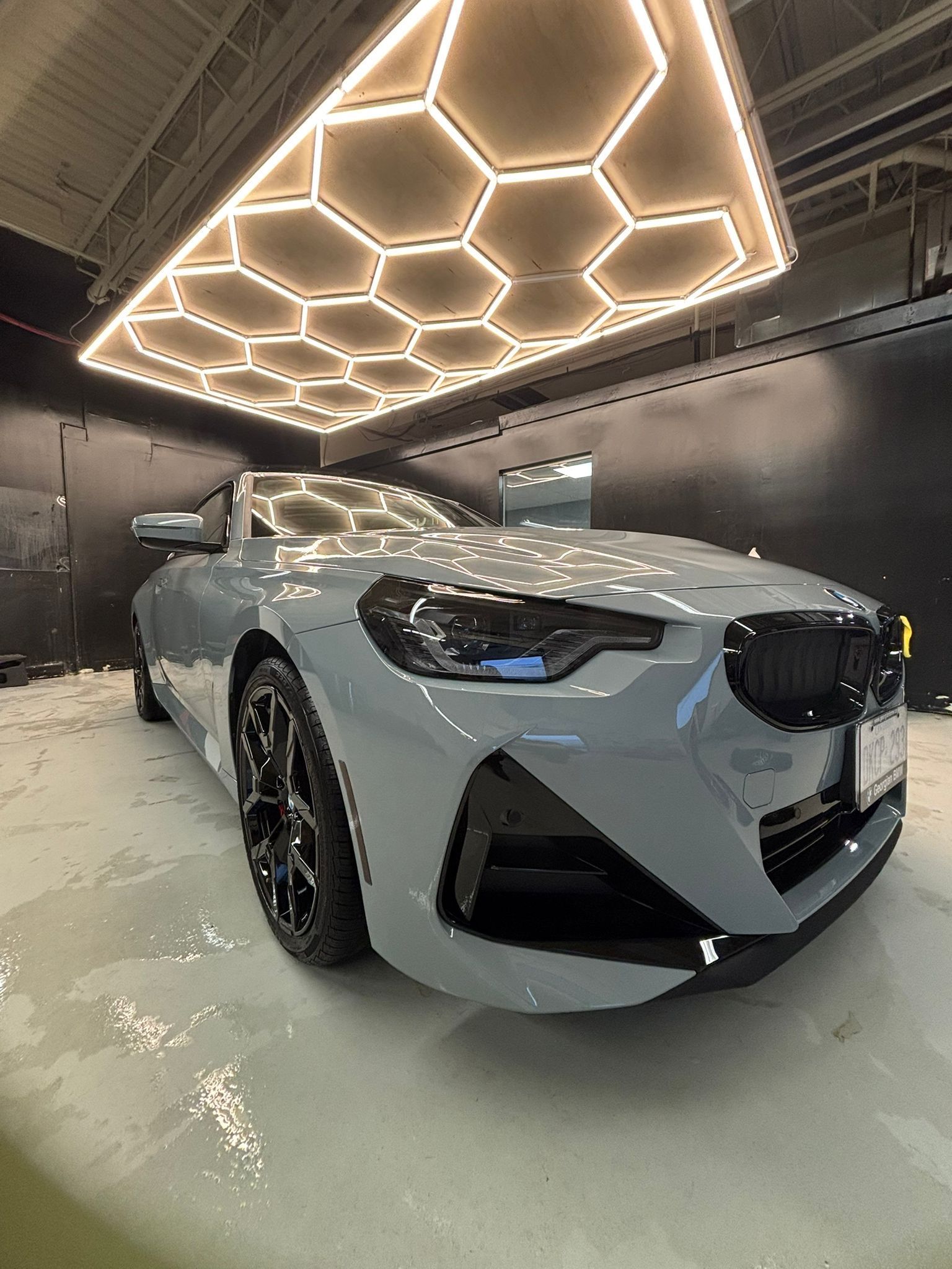 A grey BMW parked in a garage with a geometric, hexagonal LED ceiling light panel reflecting on the car's hood.