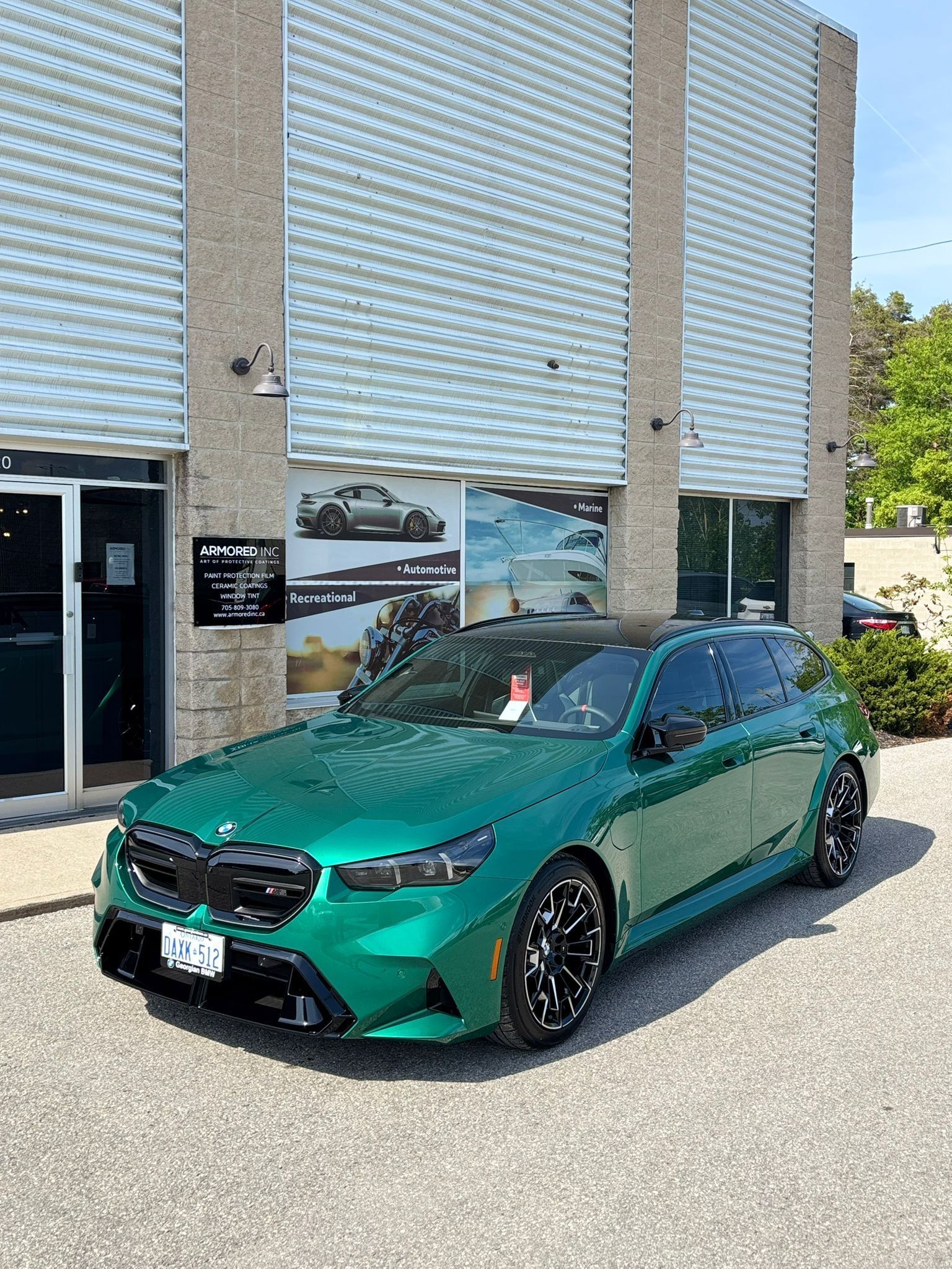 A metallic green BMW M5 Touring wagon parked on a gravel lot in front of a commercial building.