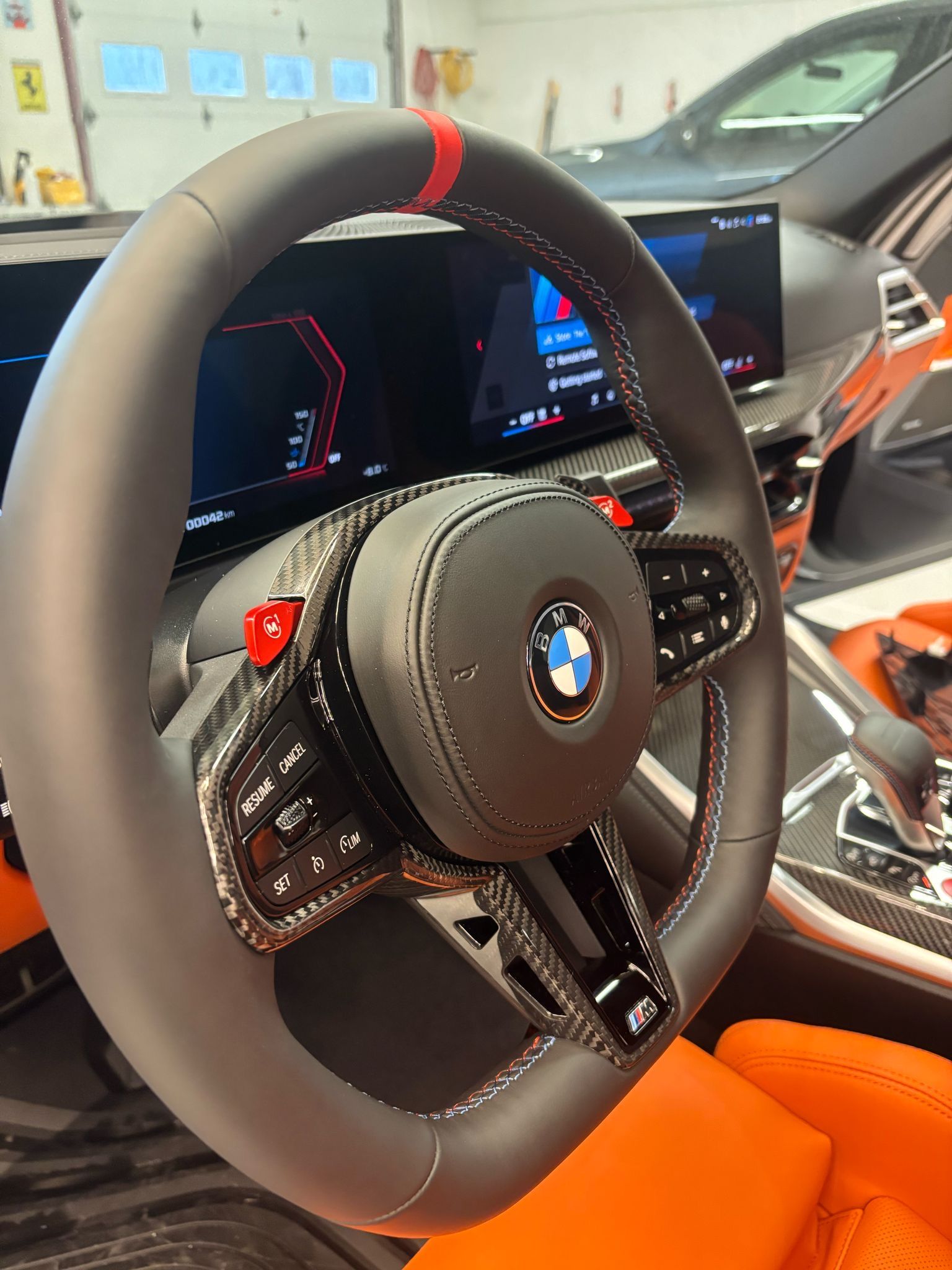 A close-up of a carbon fiber BMW steering wheel with a red center marker and buttons, set against a modern car interior.
