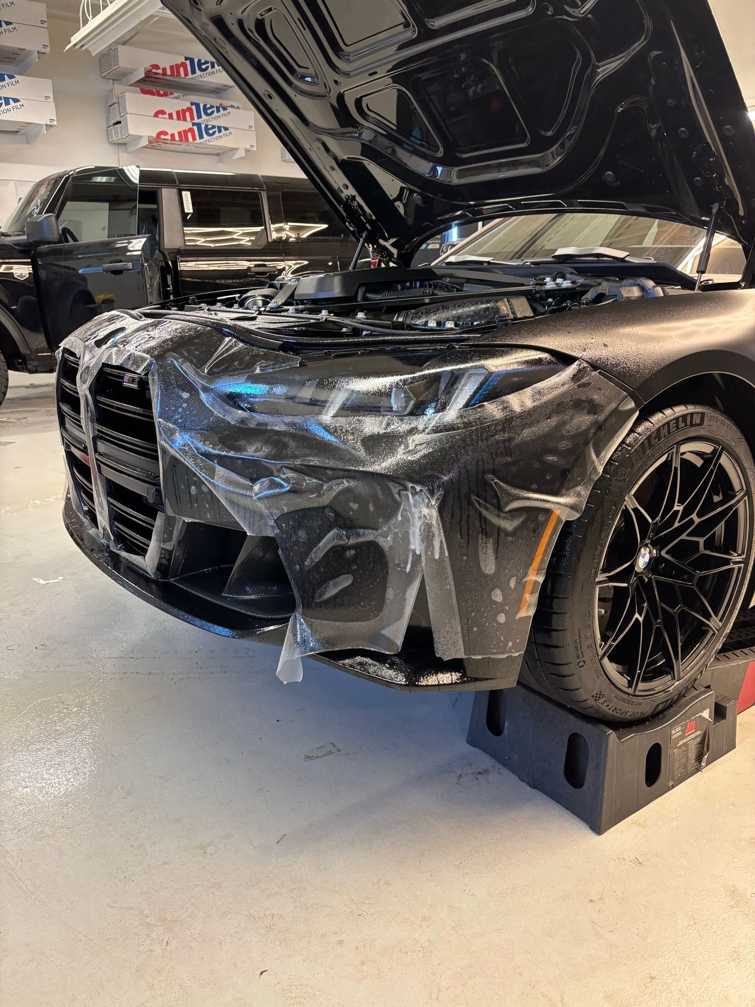 A black BMW M3 with its hood open, undergoing paint protection film installation in a garage.