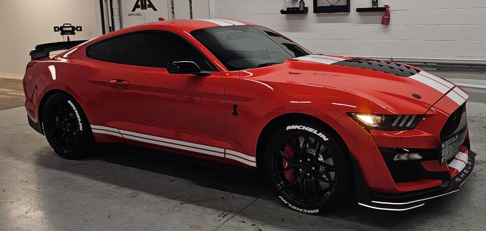 Red Ford Mustang Shelby GT500 with white racing stripes, black rims, and a rear spoiler, parked in a garage.