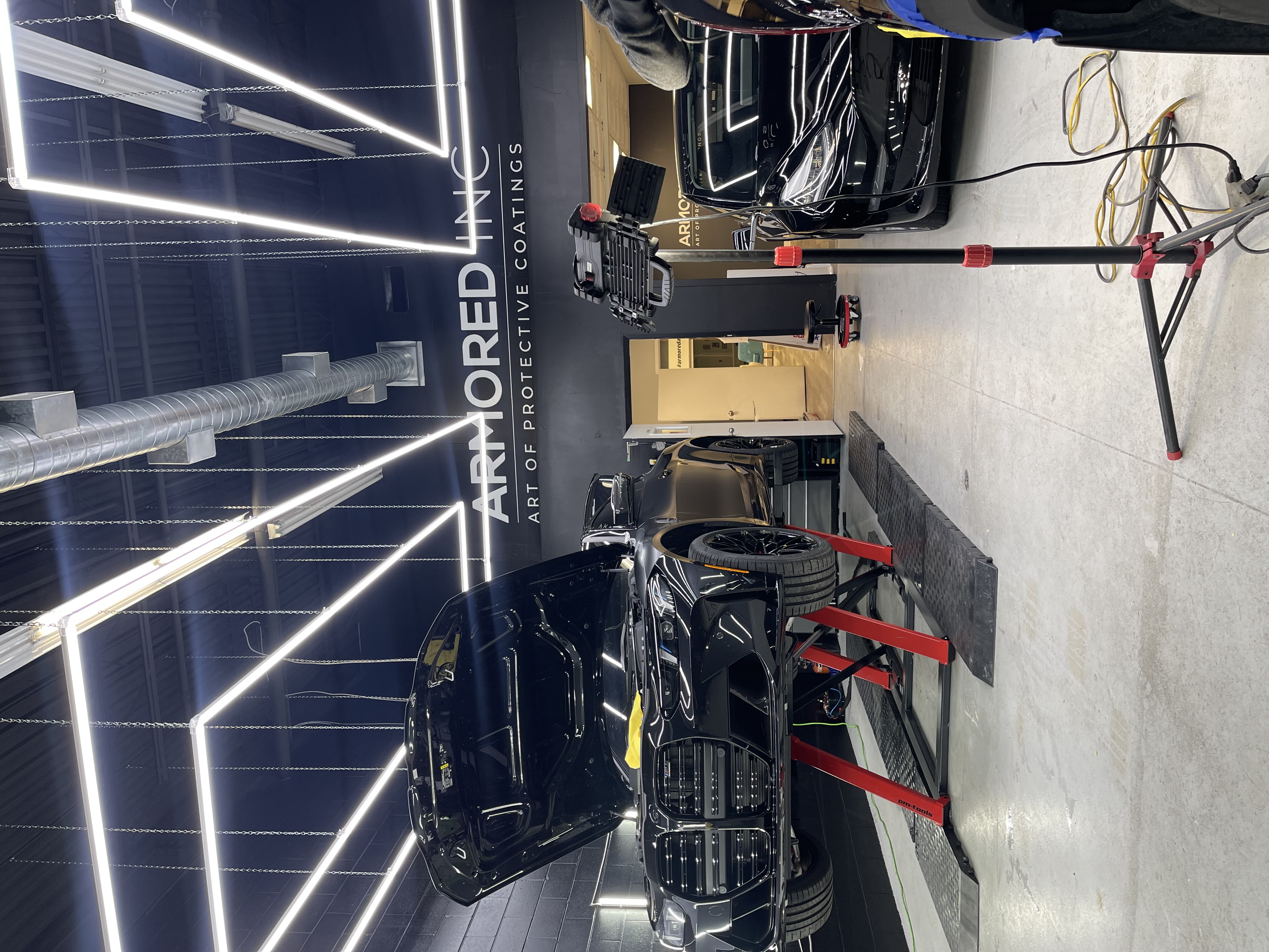 Black car on a lift in a workshop with overhead LED lighting; another car is parked to the right. 