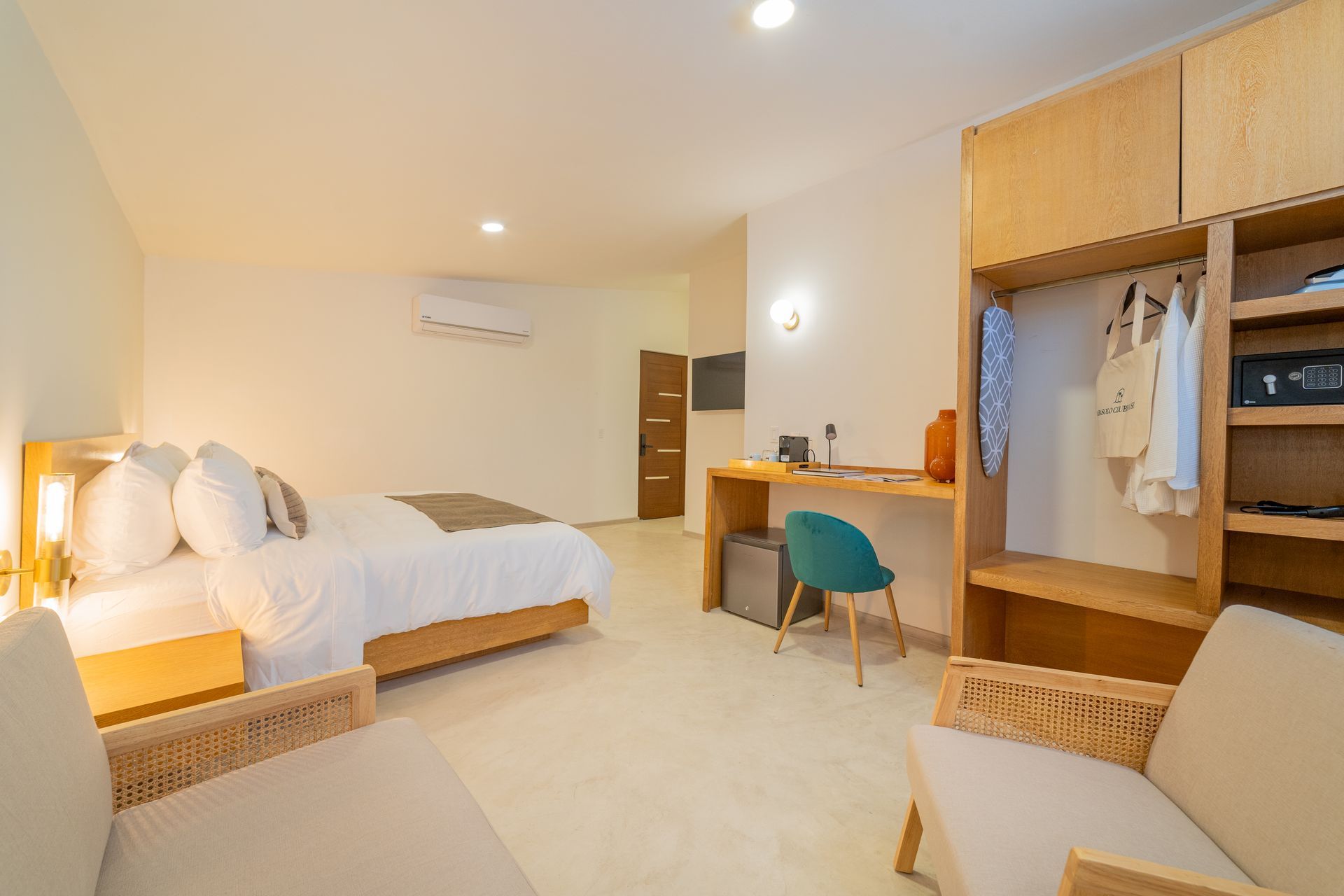 Habitación de hotel con cama, escritorio, armario y asientos. Madera clara y colores neutros.