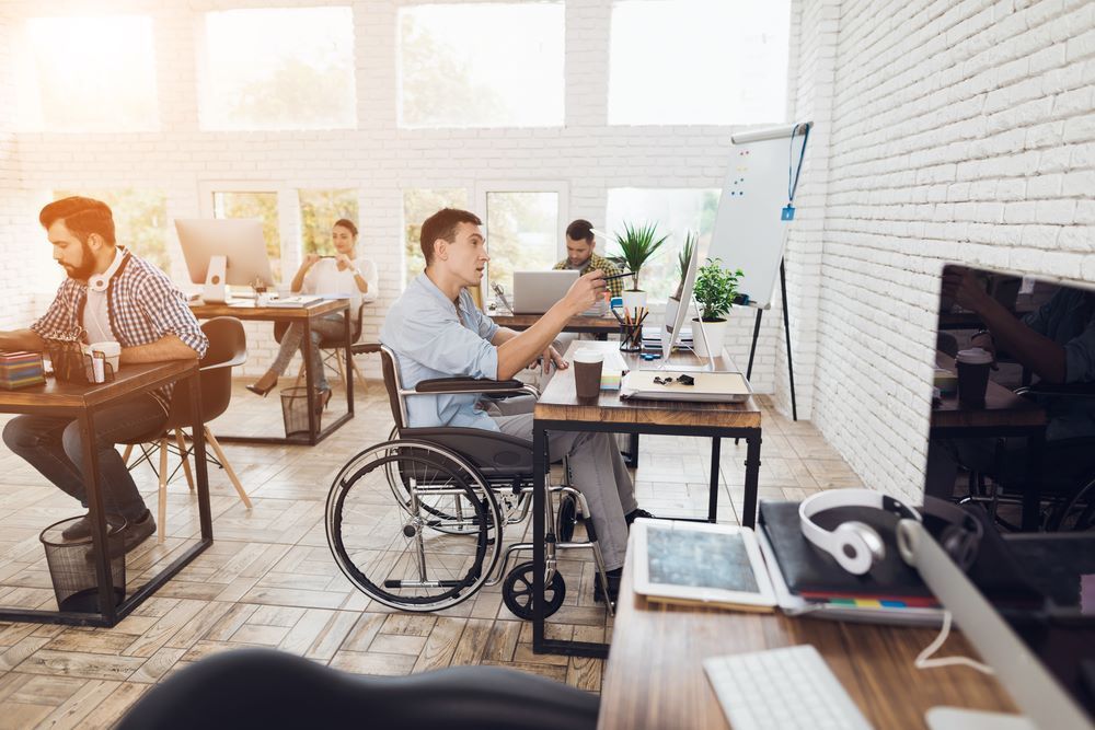 A Man in a Wheelchair is Sitting at a Desk in an Office — Supportability Care Services in West Rockhampton, QLD