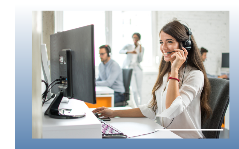 Woman wearing a headset works at a desk in a bright office, smiling at a computer.