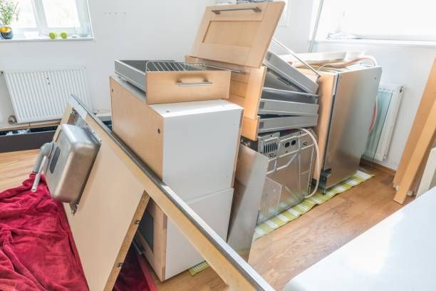 Disassembled kitchen cabinets and a stainless steel dishwasher stacked in a room with hardwood floors.