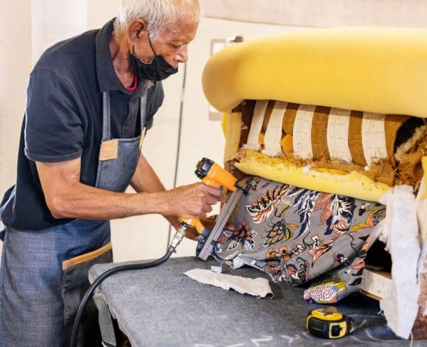 Technician using a staple gun to repair a yellow upholstered chair frame in a workshop