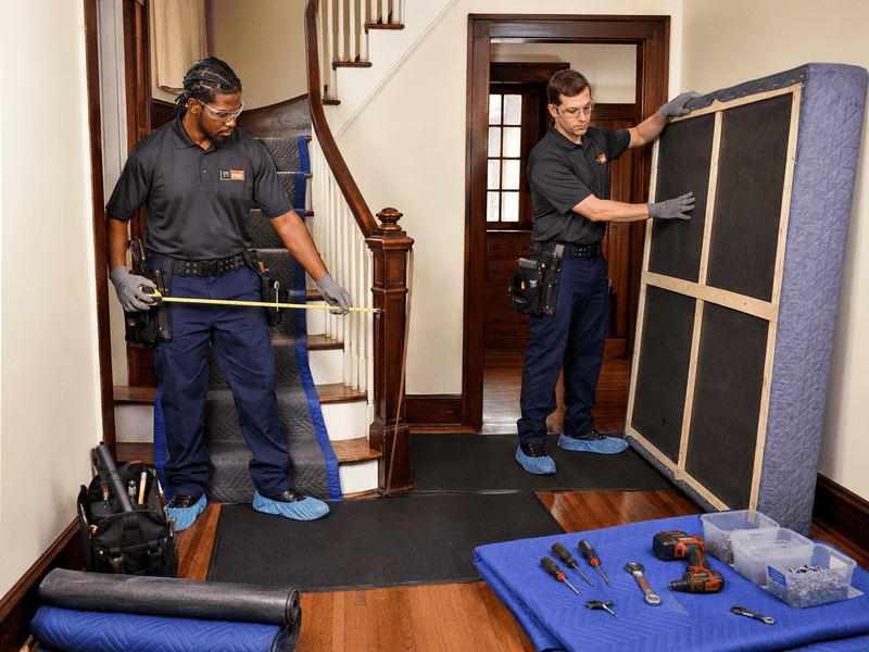 Two technicians handling large framed panels in a hallway, with tools and materials laid out on the floor.
