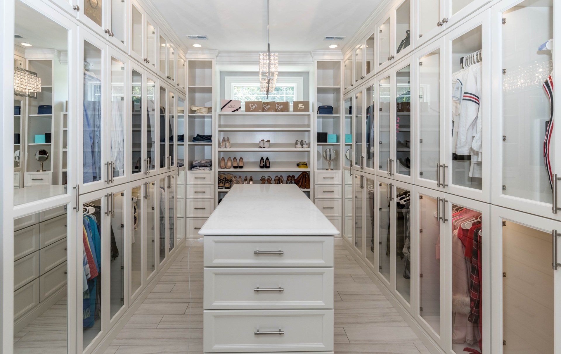 Walk-in closet with white cabinets, glass doors, and a central island. Clothes and accessories are visible.
