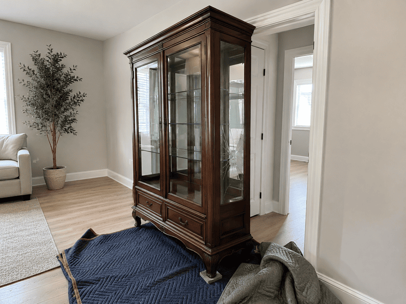 Tall wooden glass display cabinet in a bright living room, with blankets on the floor nearby.