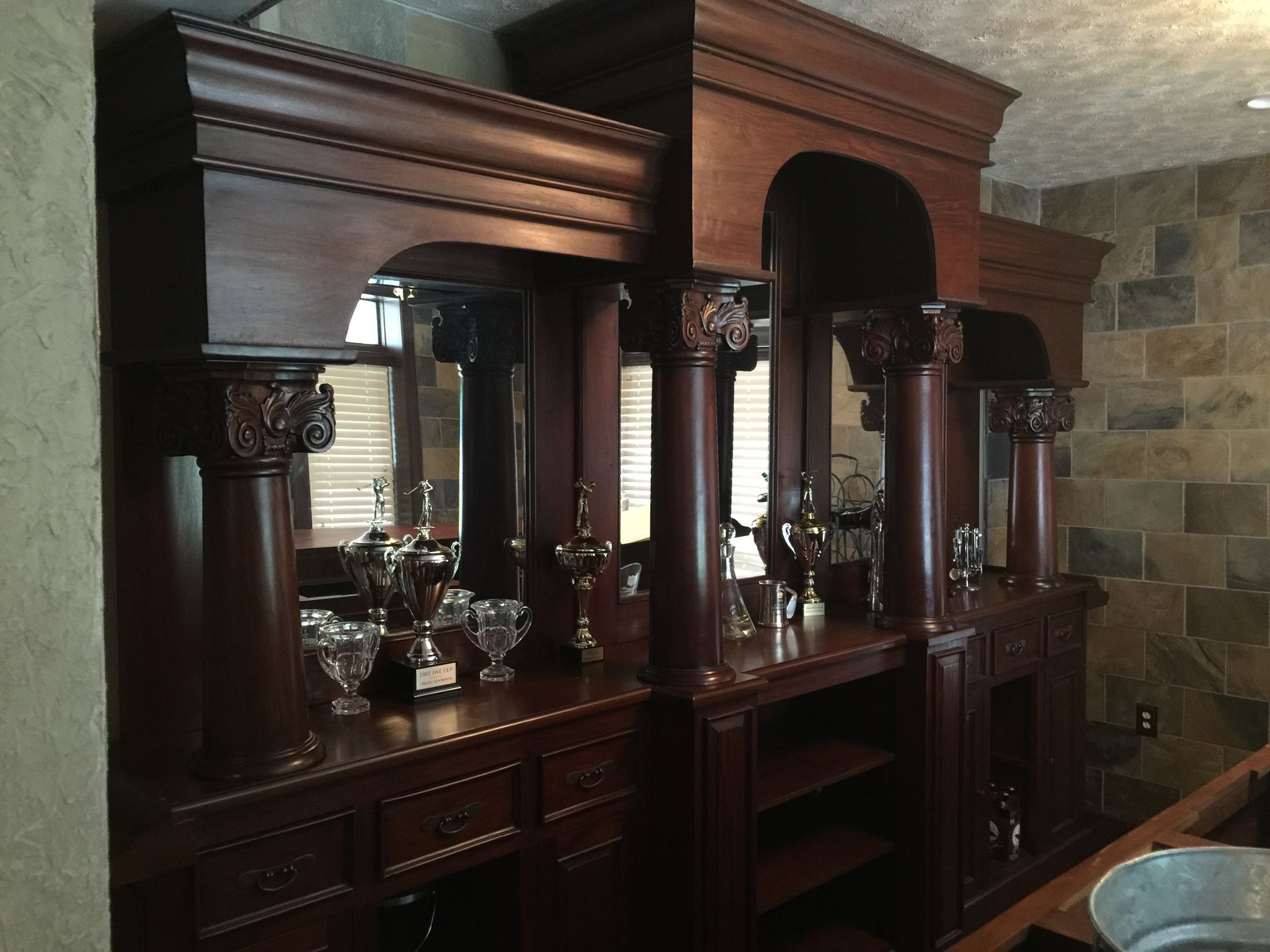 Dark wooden bar with decorative columns, mirrors, shelves, and glassware against a stone wall.