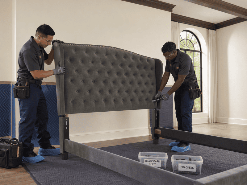 Two workers installing a gray tufted headboard in a bedroom with a bed frame and boxes nearby