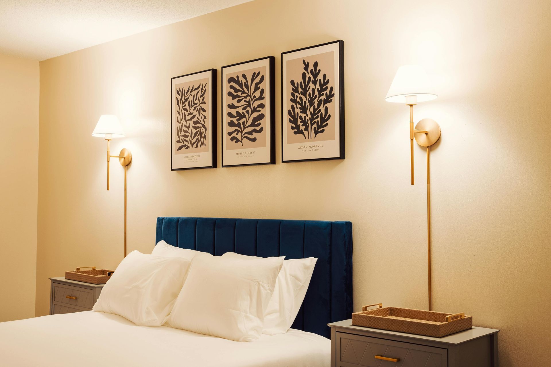 A bedroom with a blue tufted headboard, three framed botanical prints, and two gold wall-mounted lamps on beige walls.