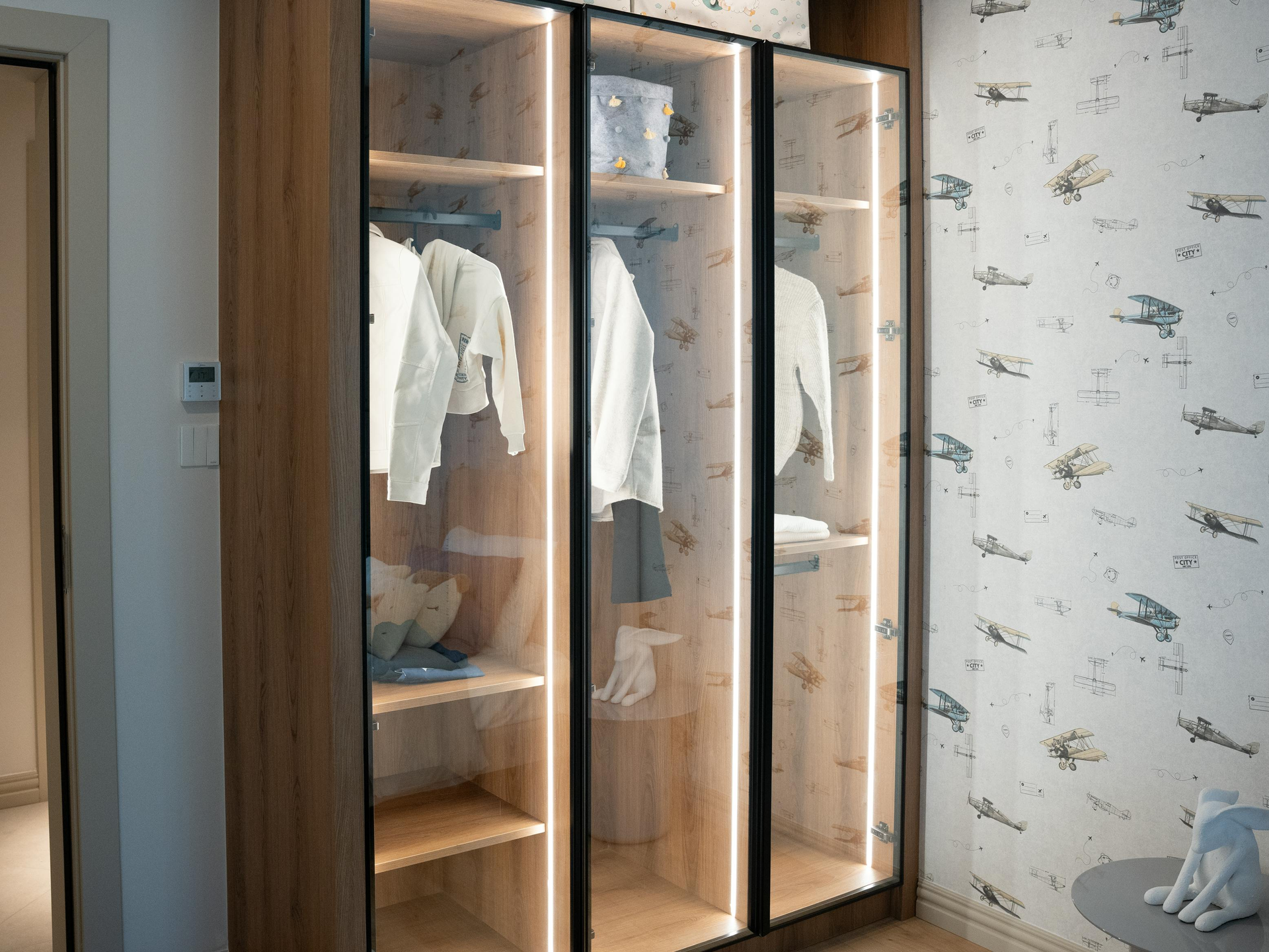 Wooden wardrobe with glass doors, illuminated interior, clothing hanging inside, set against airplane-patterned wallpaper.