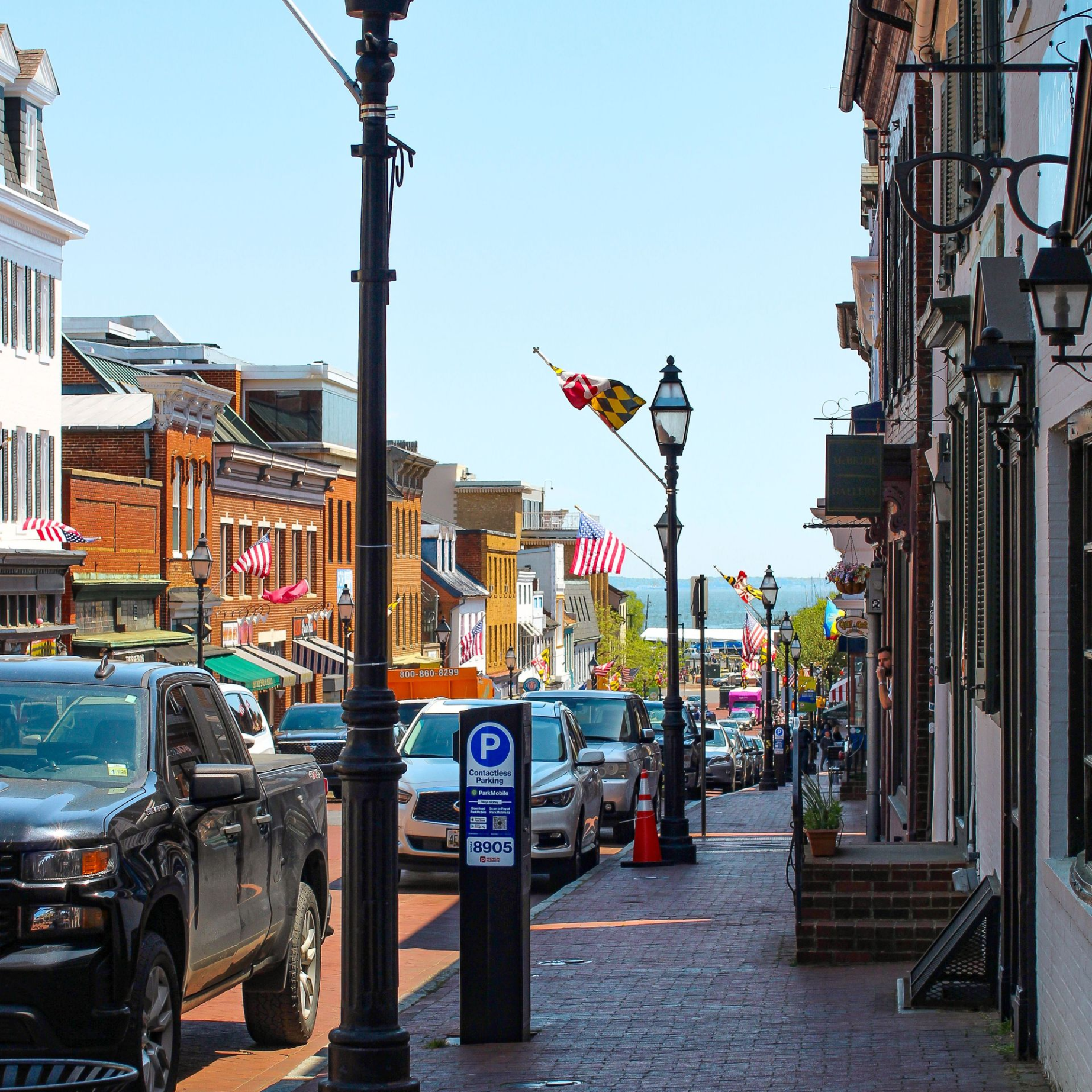 Brick sidewalk street with black lampposts, parked cars, storefronts, and American flags on a sunny day.