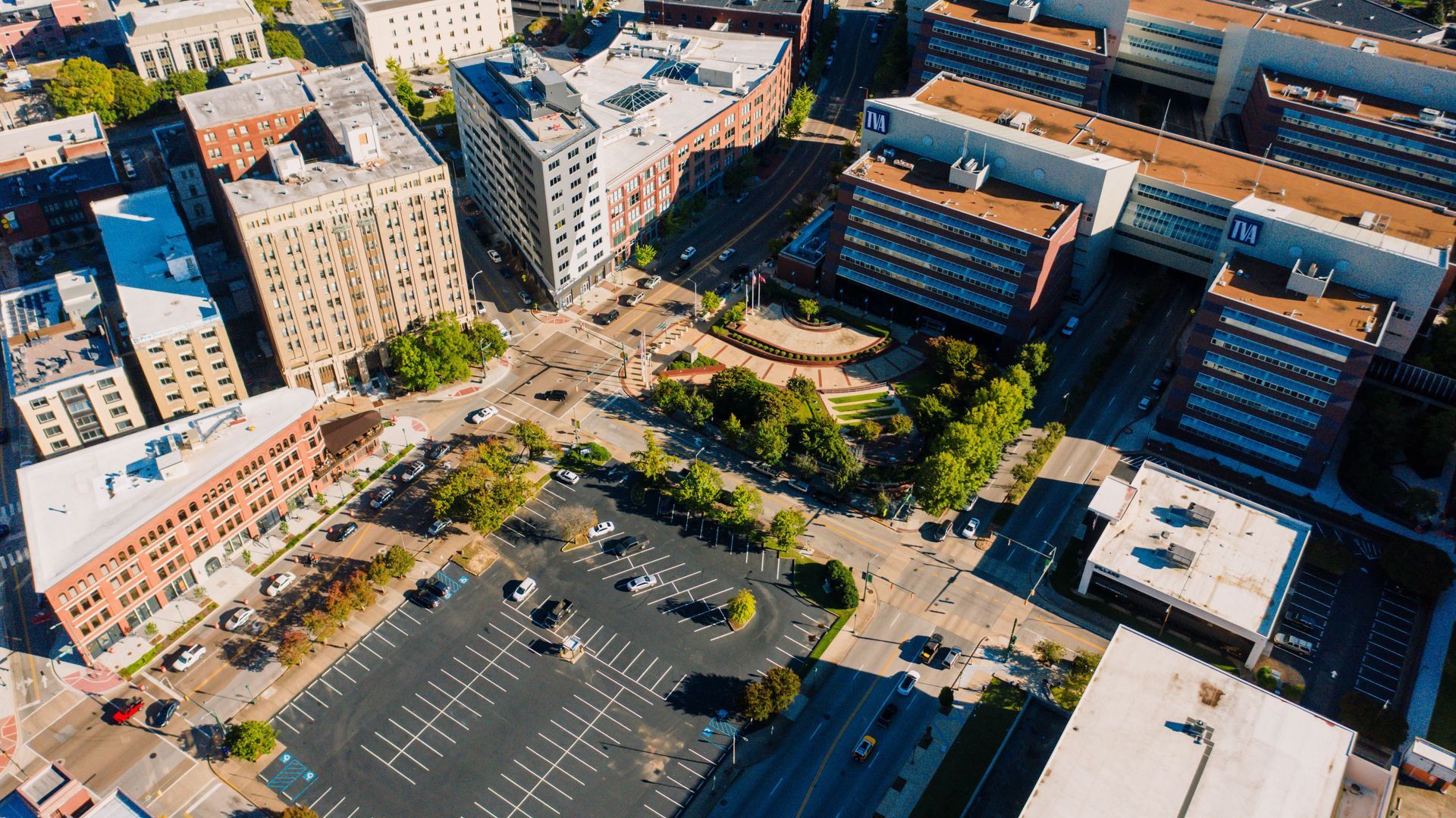 Aerial view of a city block with buildings, roads, parking lot, and a small green park.