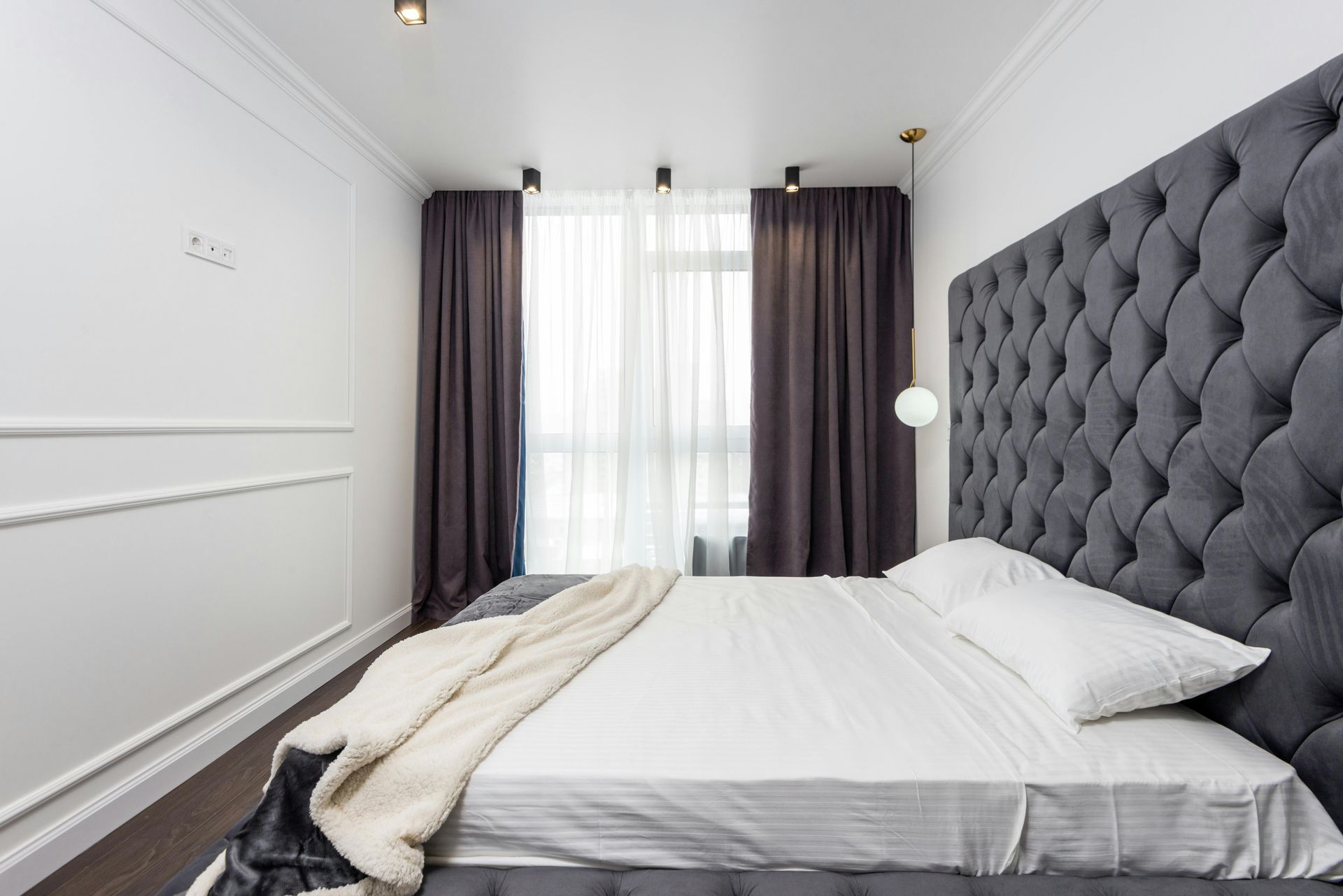 Bedroom with large gray tufted headboard, white bedding, and dark gray curtains.