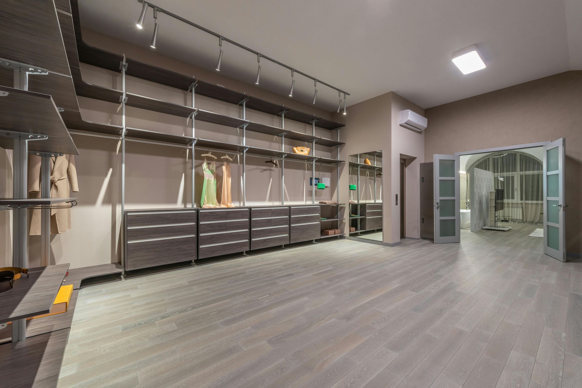 Spacious walk-in closet with gray shelves, drawers, and wood floors. Clothes are hanging. Doors lead to another room.