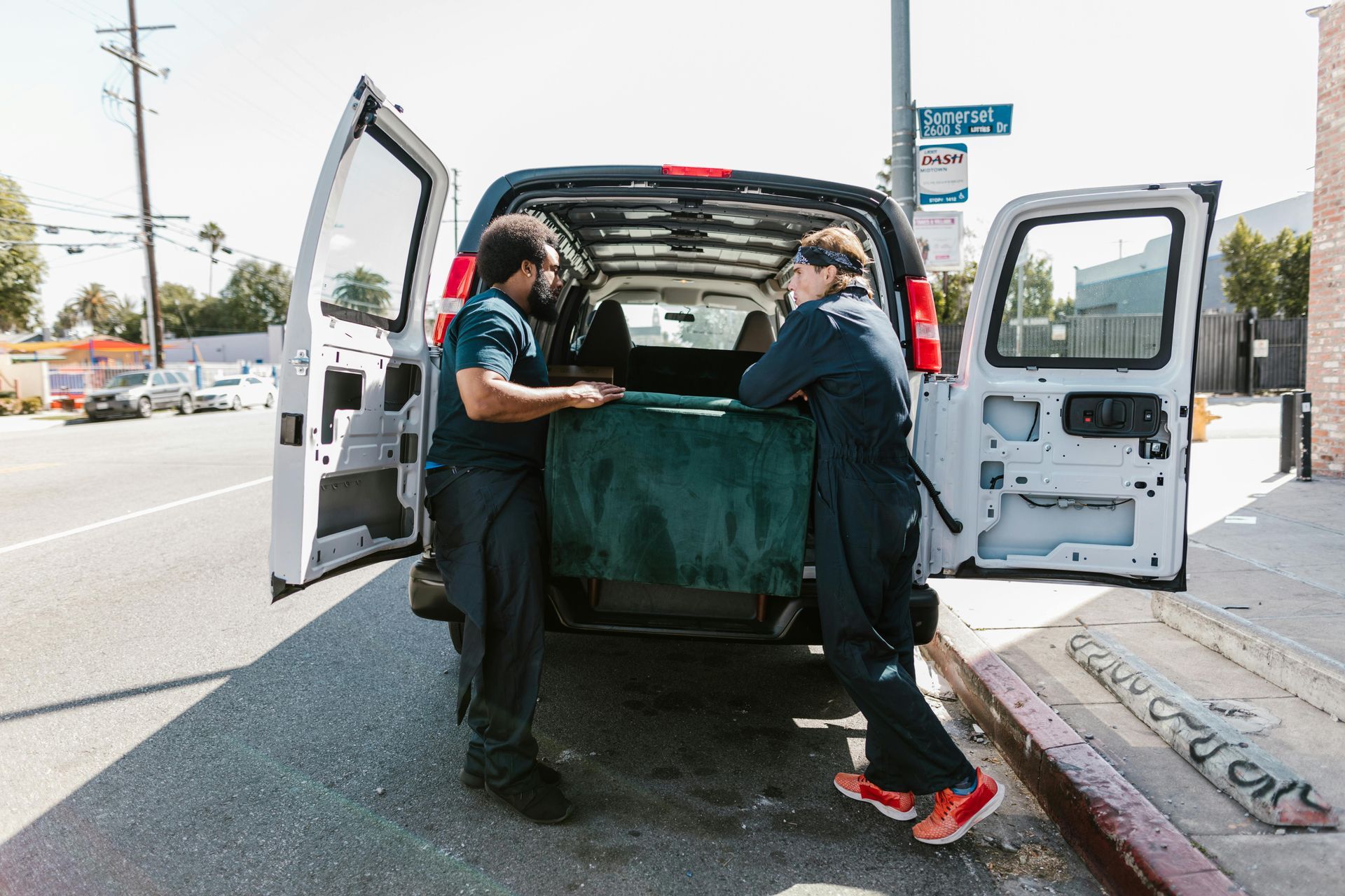 Two people in work uniforms standing by the back of an open van on a sunny city street, handling a large, covered object.