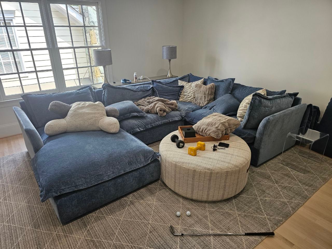 Blue sectional sofa in a bright living room with a round beige ottoman and a window beside it