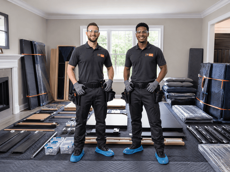 Two movers in black uniforms stand in a room filled with stacked floor mats and packed bundles.