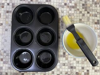 Uno stampo per muffin è appoggiato su un tavolo piastrellato, accanto a una ciotola di olio e un pennello.