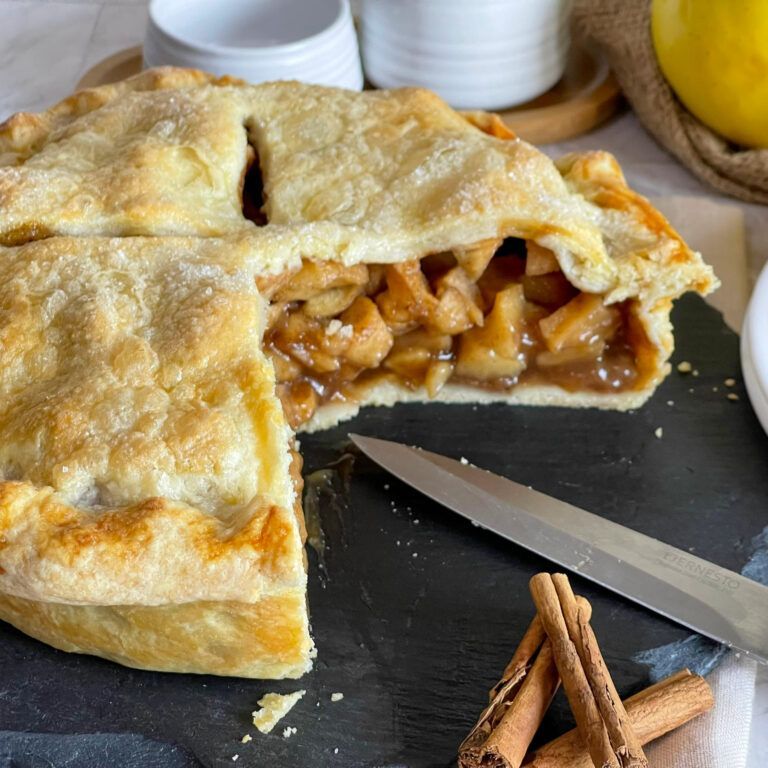 Una torta di mele con una fetta staccata si trova su un tagliere con dei bastoncini di cannella.