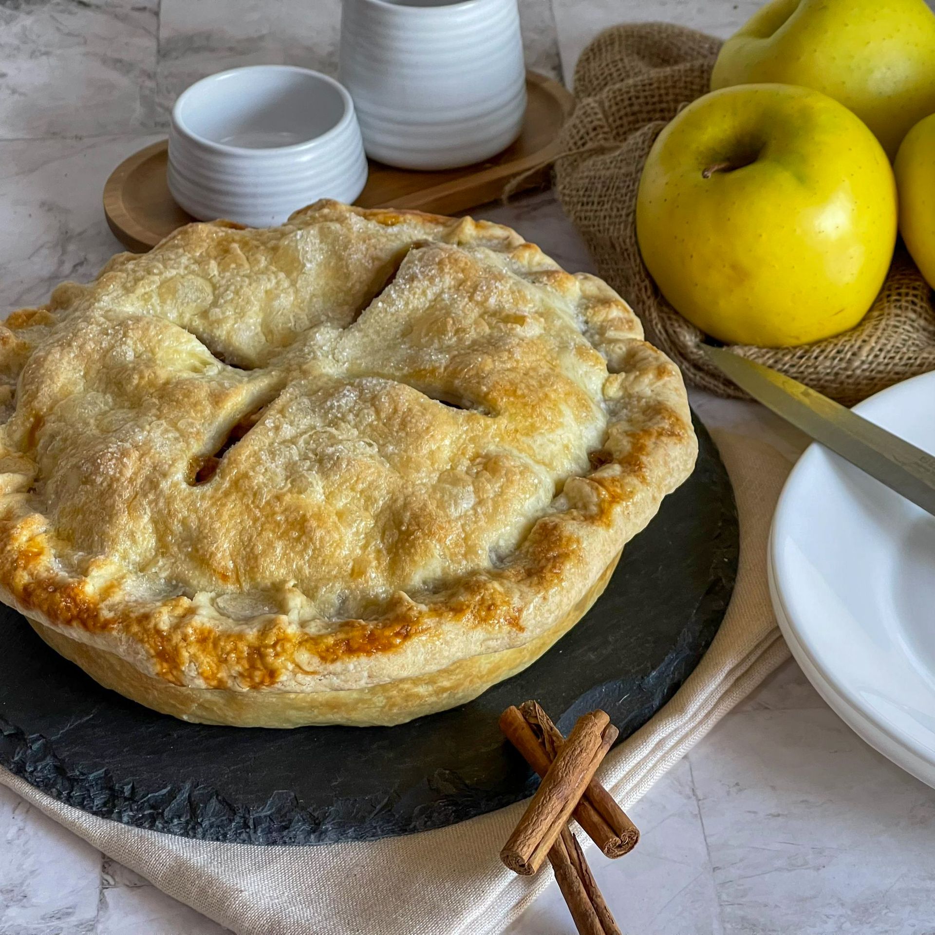 Una torta è appoggiata su un tagliere accanto a mele e bastoncini di cannella.