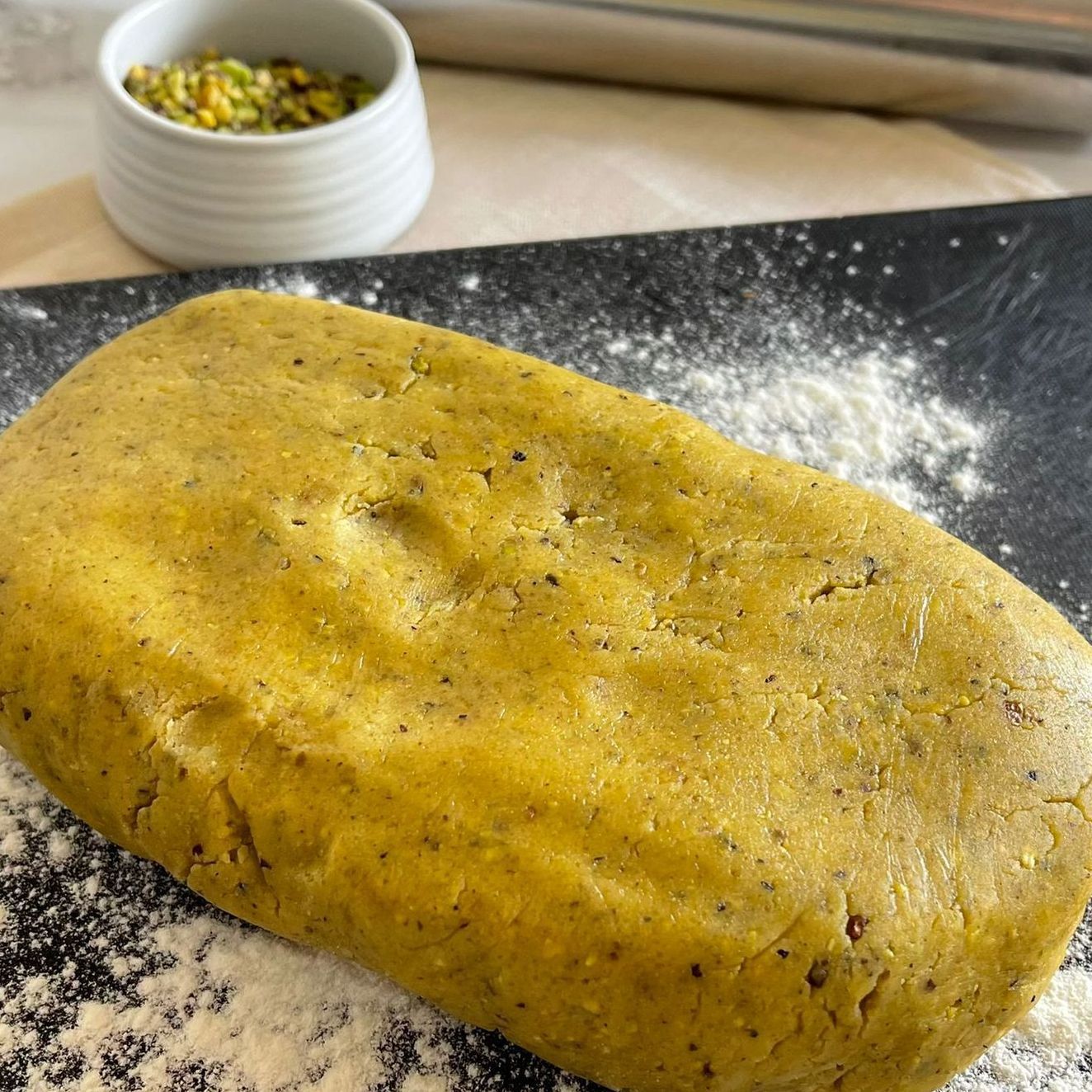 Un pezzo di pasta è appoggiato su un tagliere accanto a una ciotola di pistacchi.