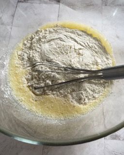 Una ciotola di vetro piena di farina e liquido con una frusta al suo interno.