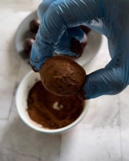 Una persona che indossa un guanto blu tiene in mano un tartufo al cioccolato sopra una ciotola di cioccolato in polvere.