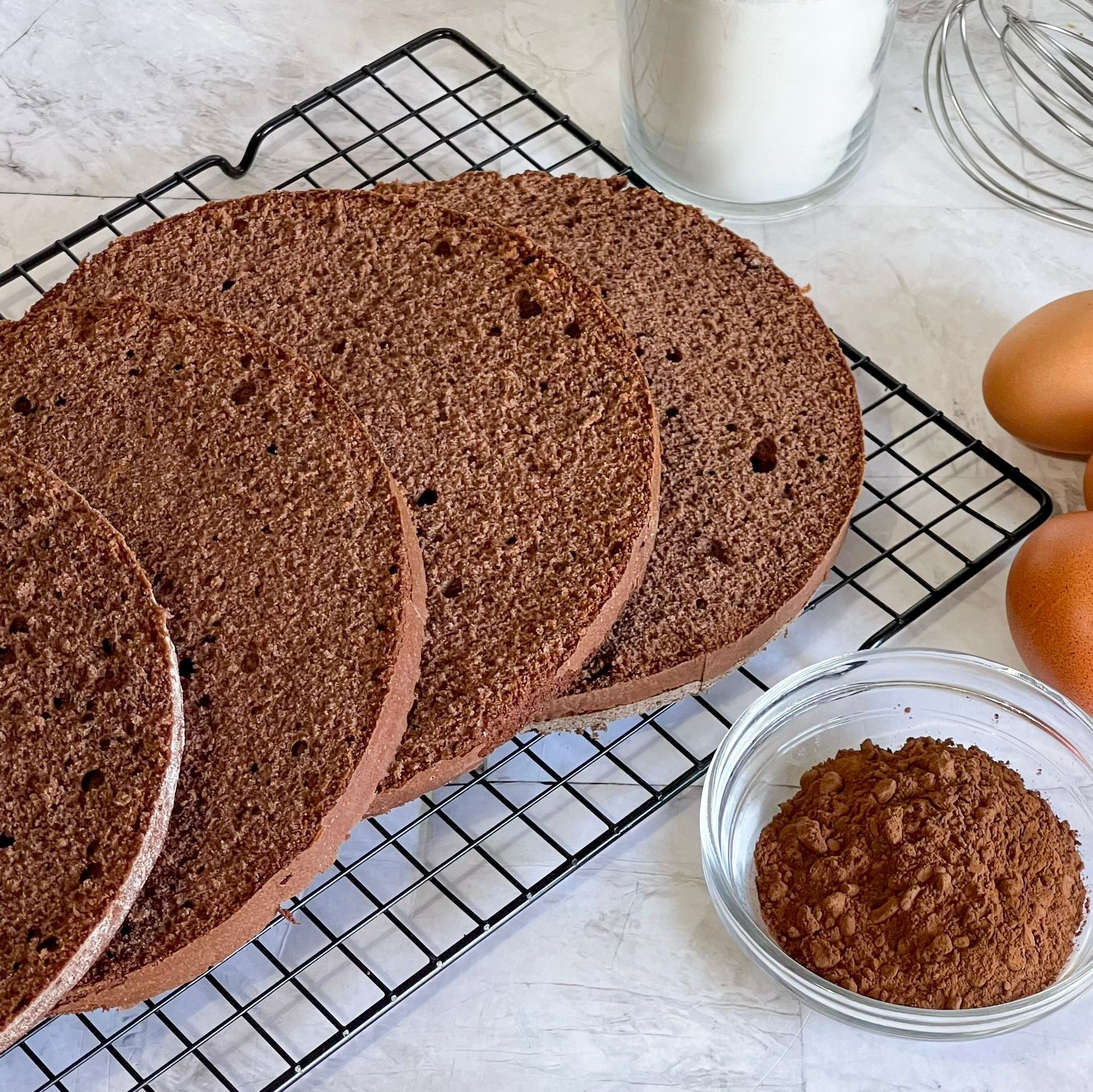 Una torta al cioccolato è appoggiata su una griglia di raffreddamento accanto a uova e cacao in polvere.