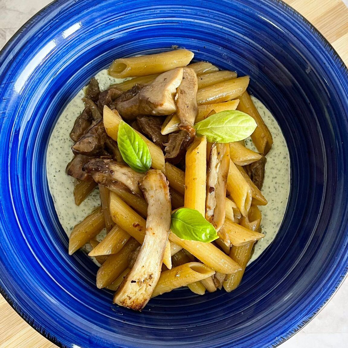Un piatto blu condito con pasta, funghi e foglie di basilico.