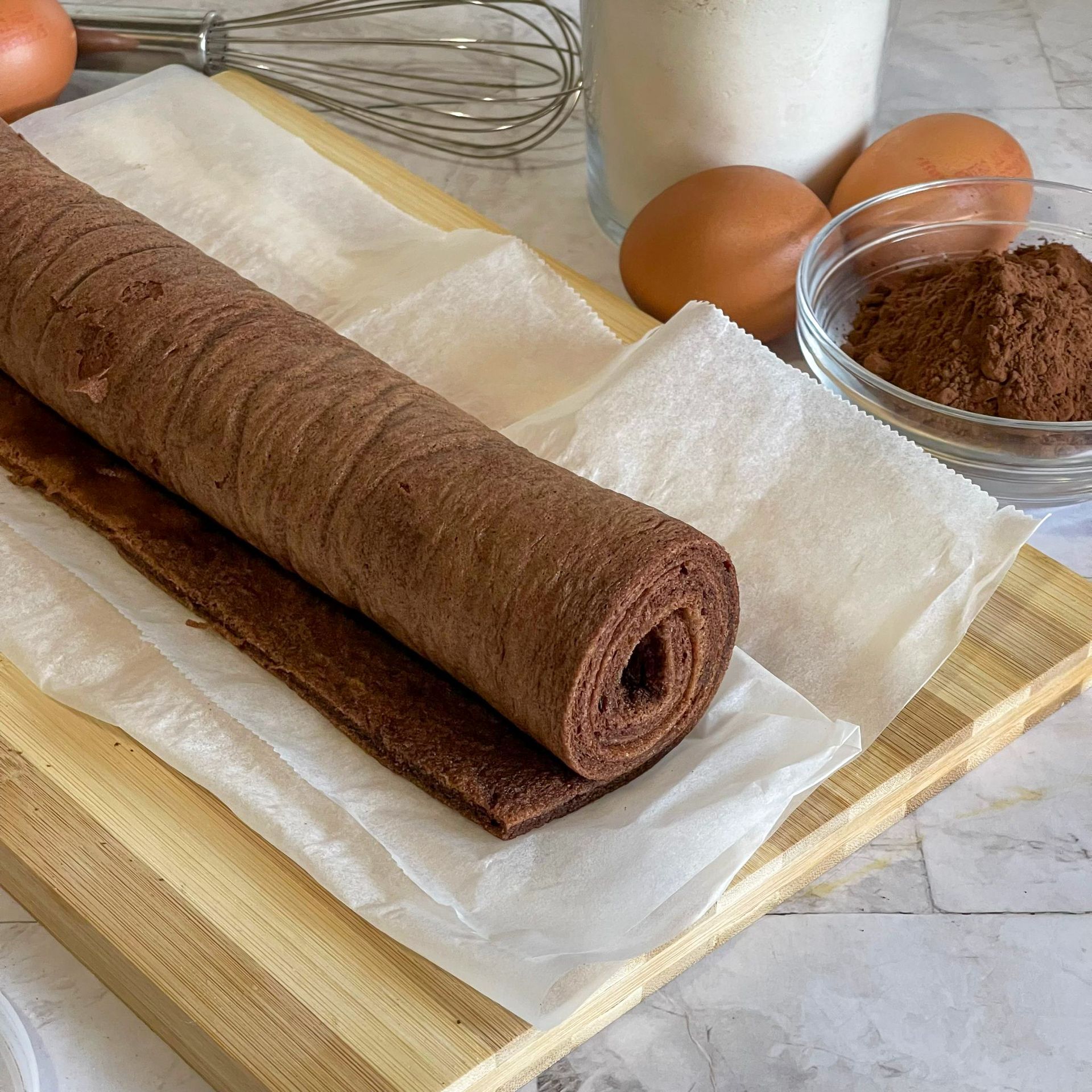 Un rotolo di cioccolato è appoggiato su un tagliere di legno accanto a uova e cacao in polvere.