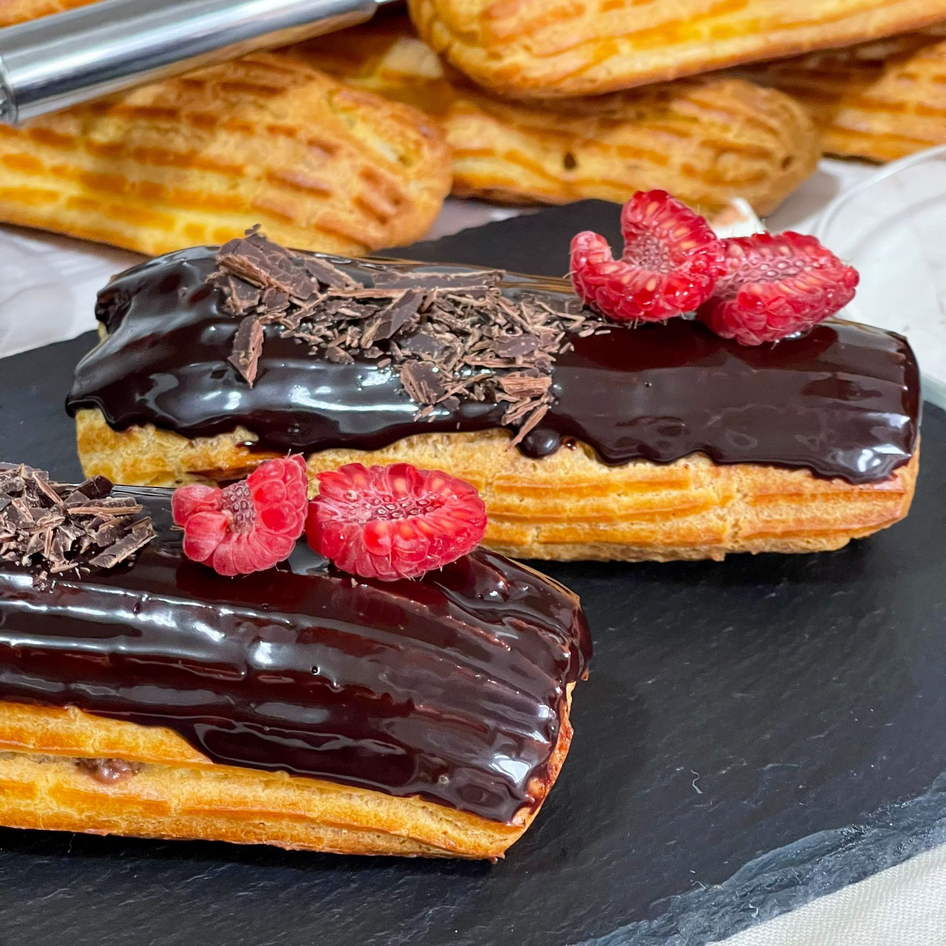 Due éclair ricoperti di cioccolato con lamponi sopra su un tagliere.