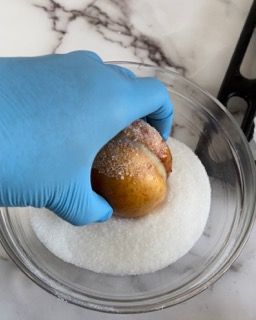 Una persona che indossa guanti blu tiene in mano una ciambella in una ciotola di zucchero.
