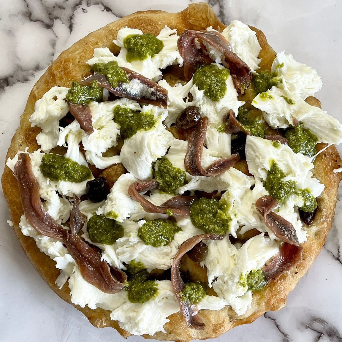 Una pizza con formaggio, pesto e acciughe su una superficie di marmo.