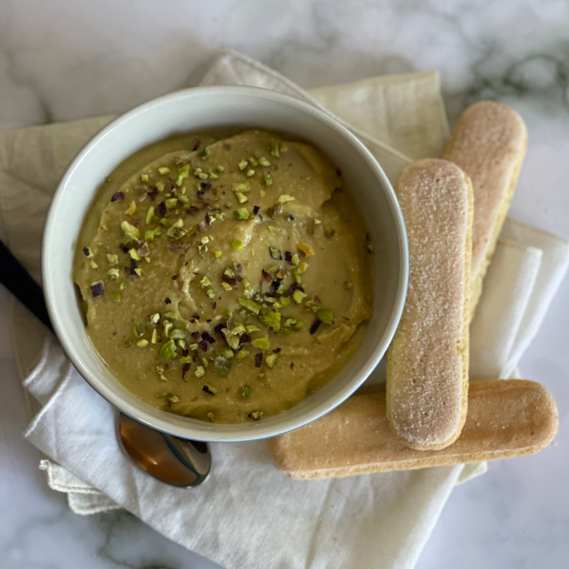 Una ciotola di zuppa con pistacchi sopra accanto a due bastoncini di tiramisù e un cucchiaio.