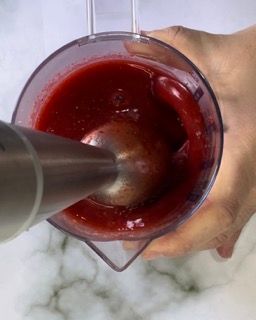 Una persona sta usando un frullatore per mescolare un liquido rosso in un misurino.